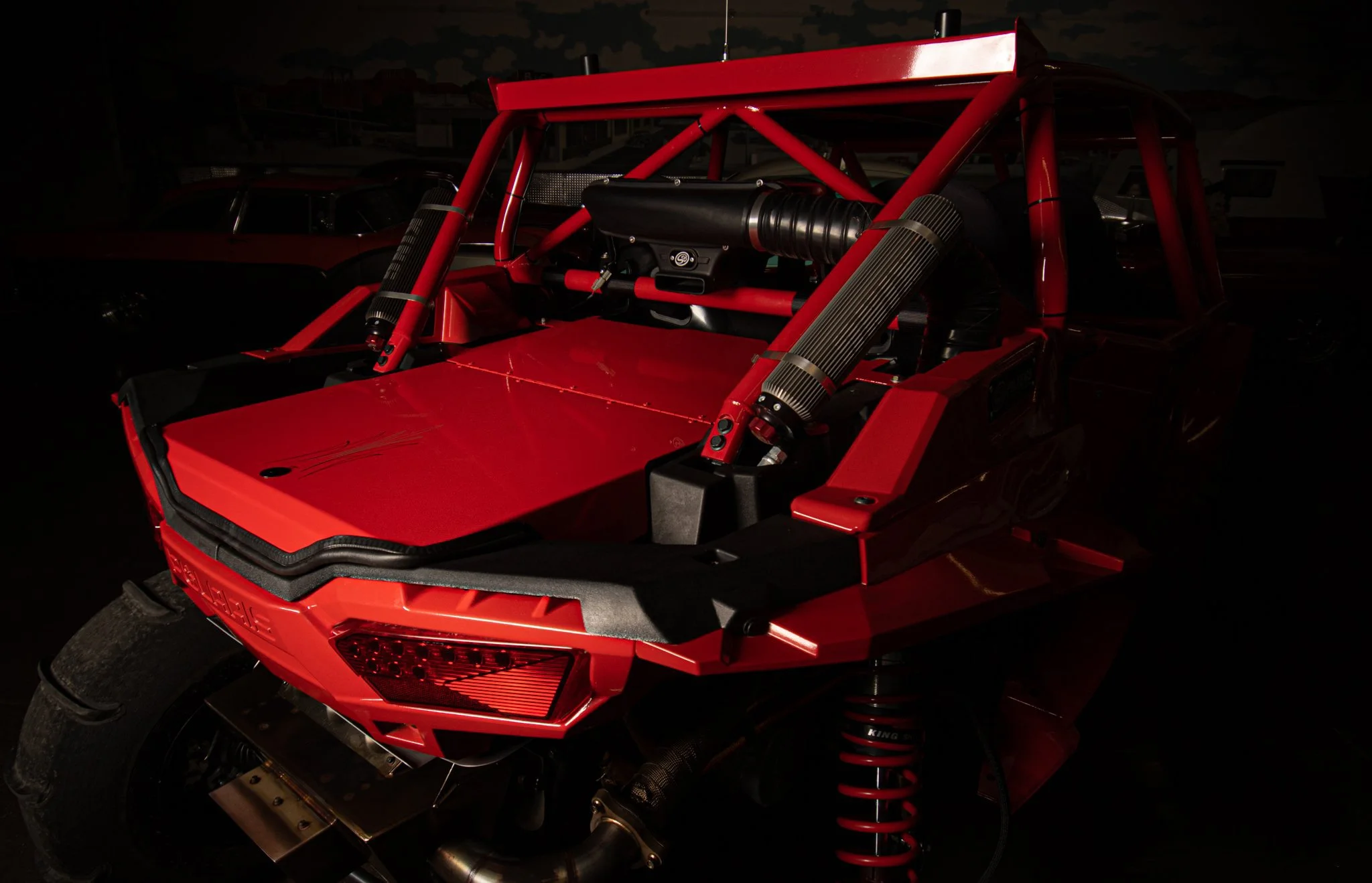 A red race car without doors or windows displaying its internal structure, including the engine, suspension, and roll cage, on display in a dimly lit setting.