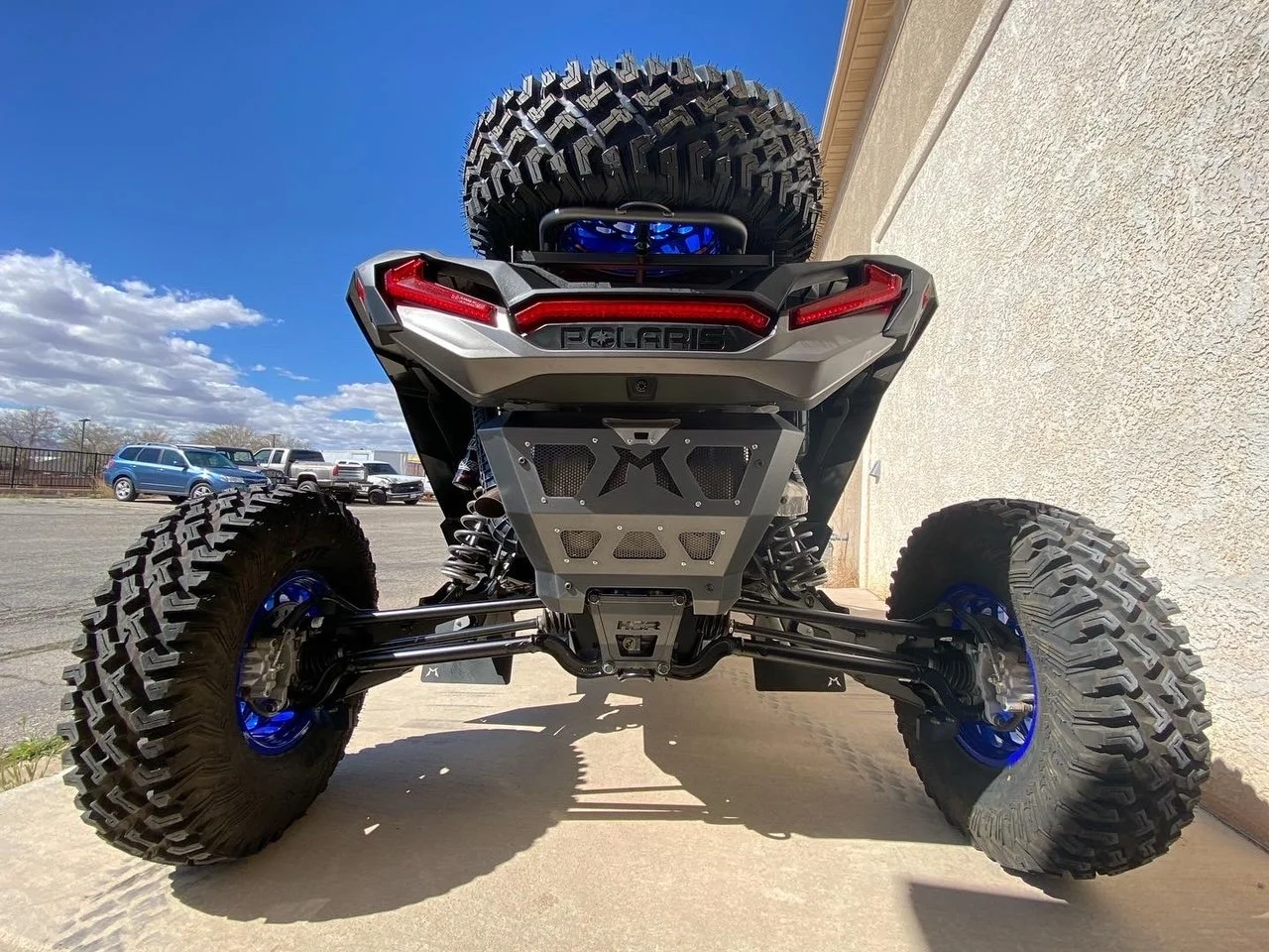 From the rear view, a Polaris off-road vehicle with large tires, a spare tire on the back, and a silver and black body with red accents, parked on a concrete surface near a building under a partly cloudy sky.