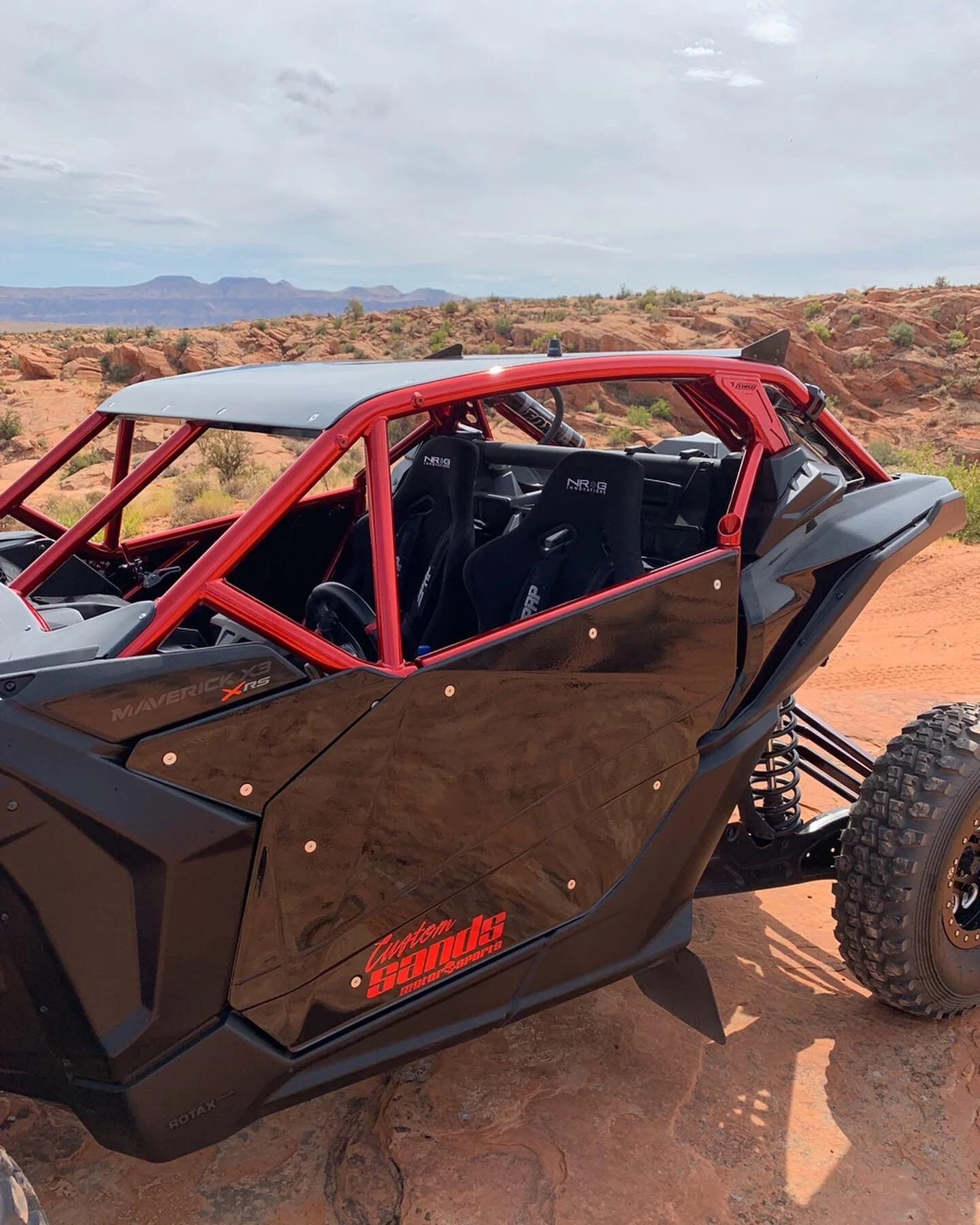 Off-road dune buggy with black body and red roll cage on desert terrain with rocky landscape and mountains in the background.