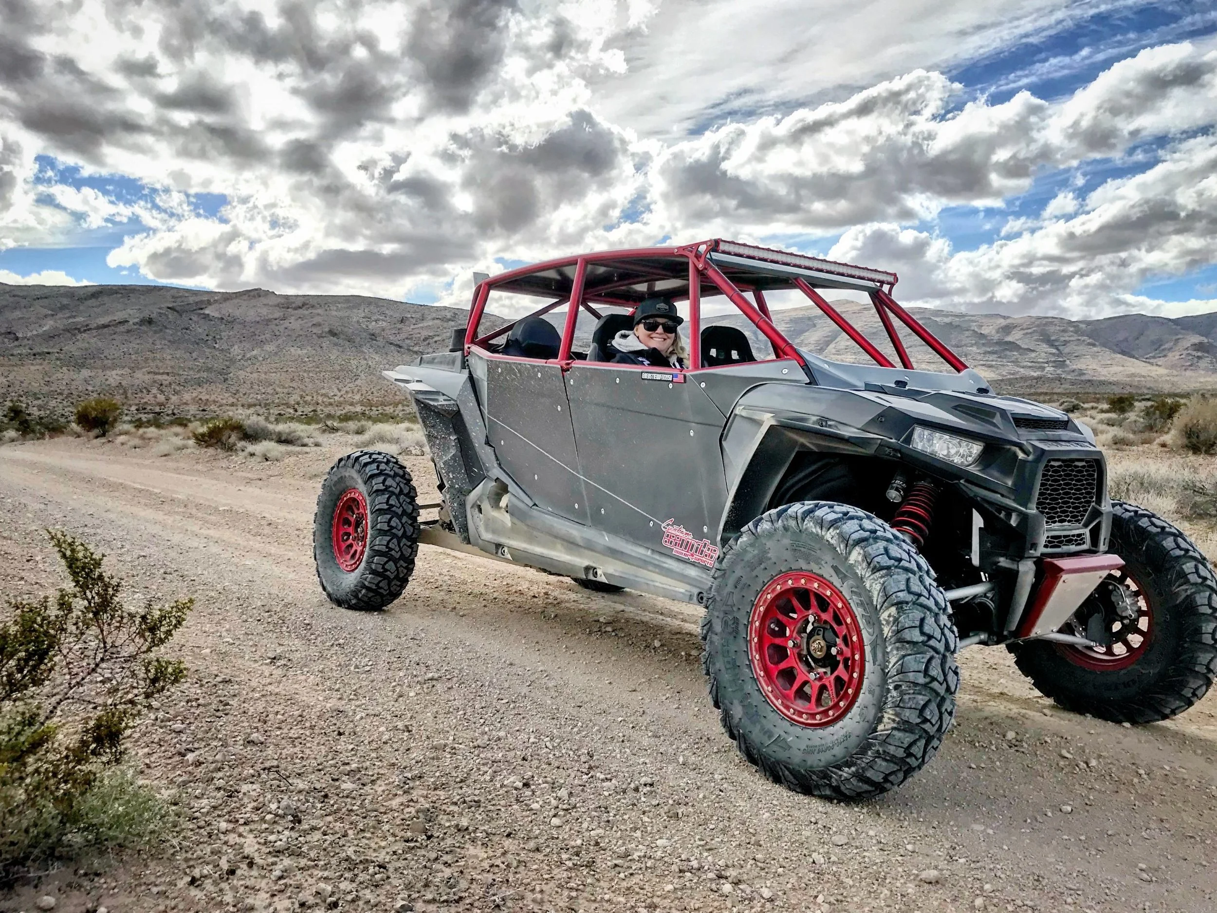 A woman sitting in a black off-road vehicle with red roll bars on a dirt road in a desert landscape, with mountains and a sky filled with scattered clouds in the background.