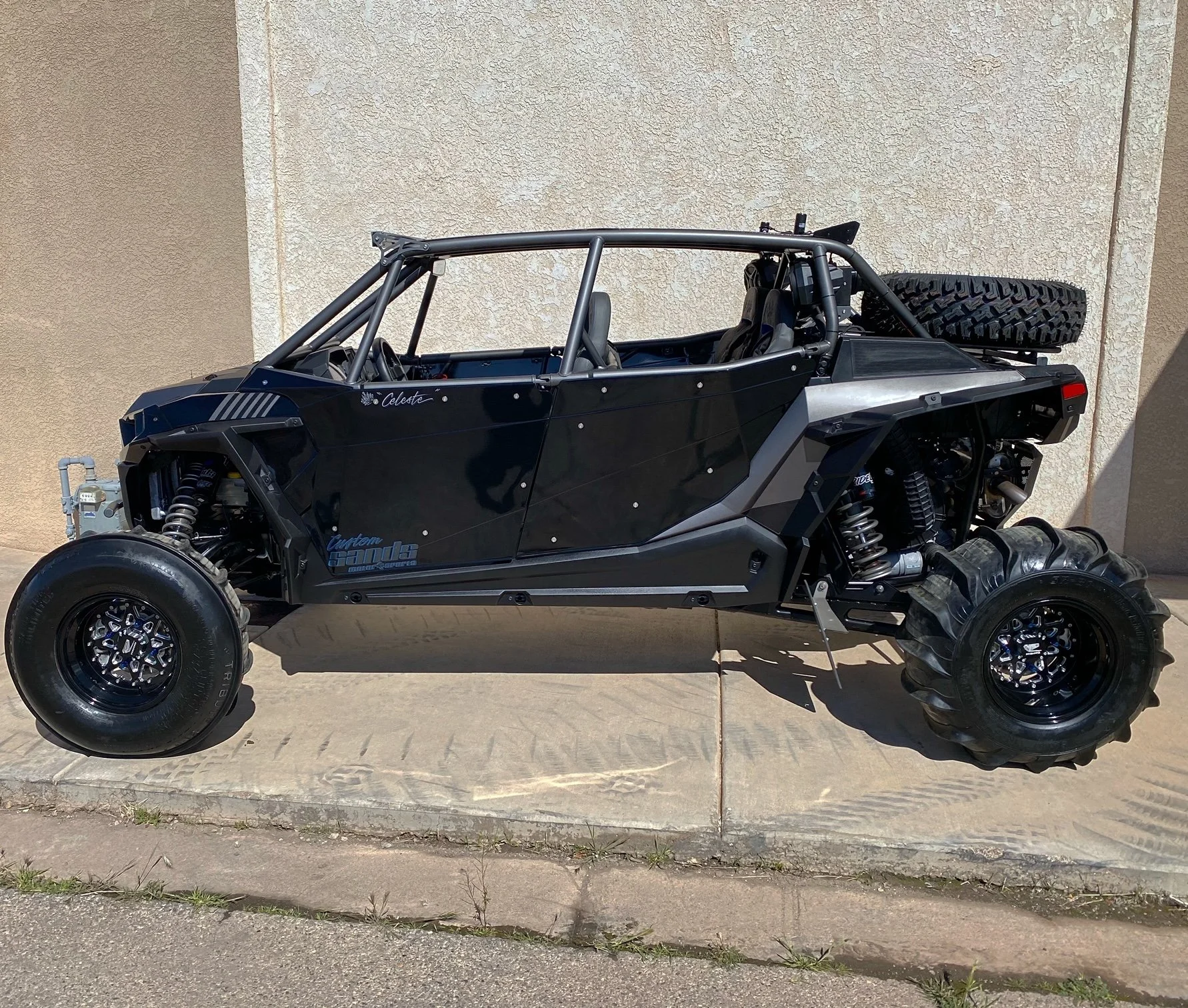 A custom-built off-road or dune buggy with large tires, black body, and tubular frame, parked against a beige wall.
