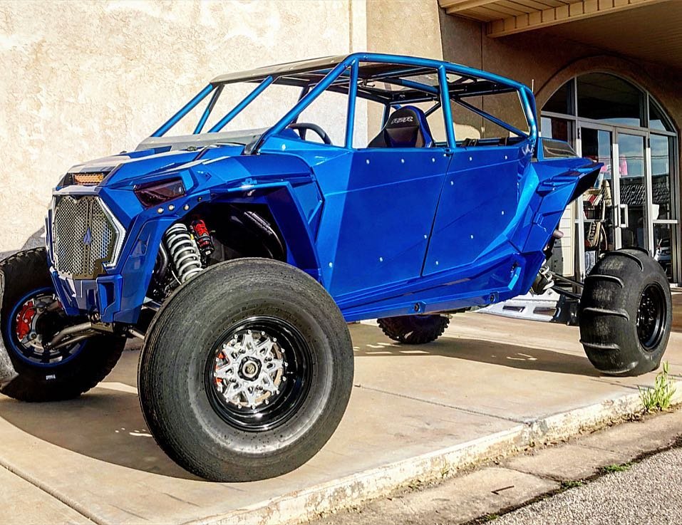 A custom blue off-road dune buggy with large rear tires and exposed suspension, parked outside a building with large glass doors.