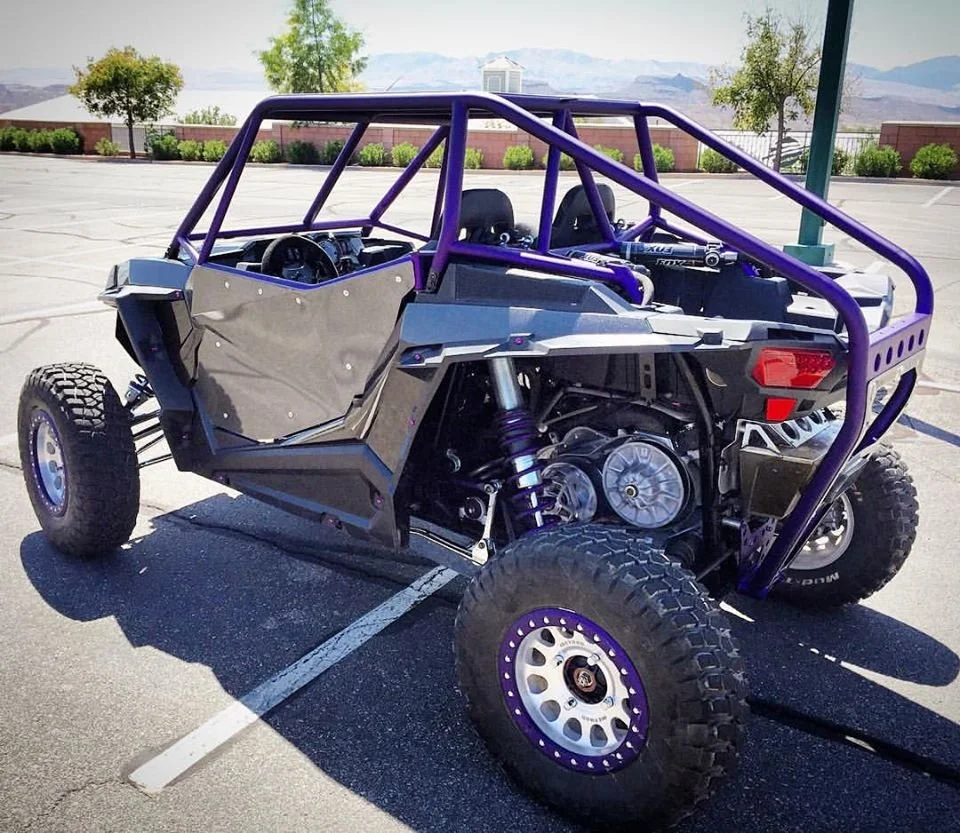 An off-road racing dune buggy parked in an empty parking lot with mountains and trees in the background.
