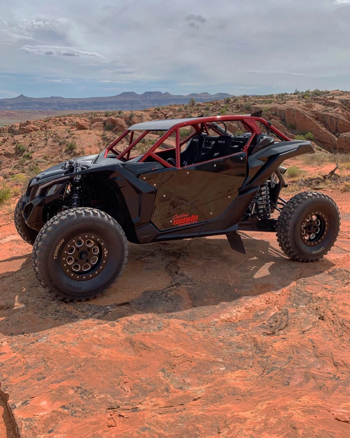 An off-road dune buggy vehicle with a black body and red frame, parked on rocky desert terrain with mountains and cloudy sky in the background.