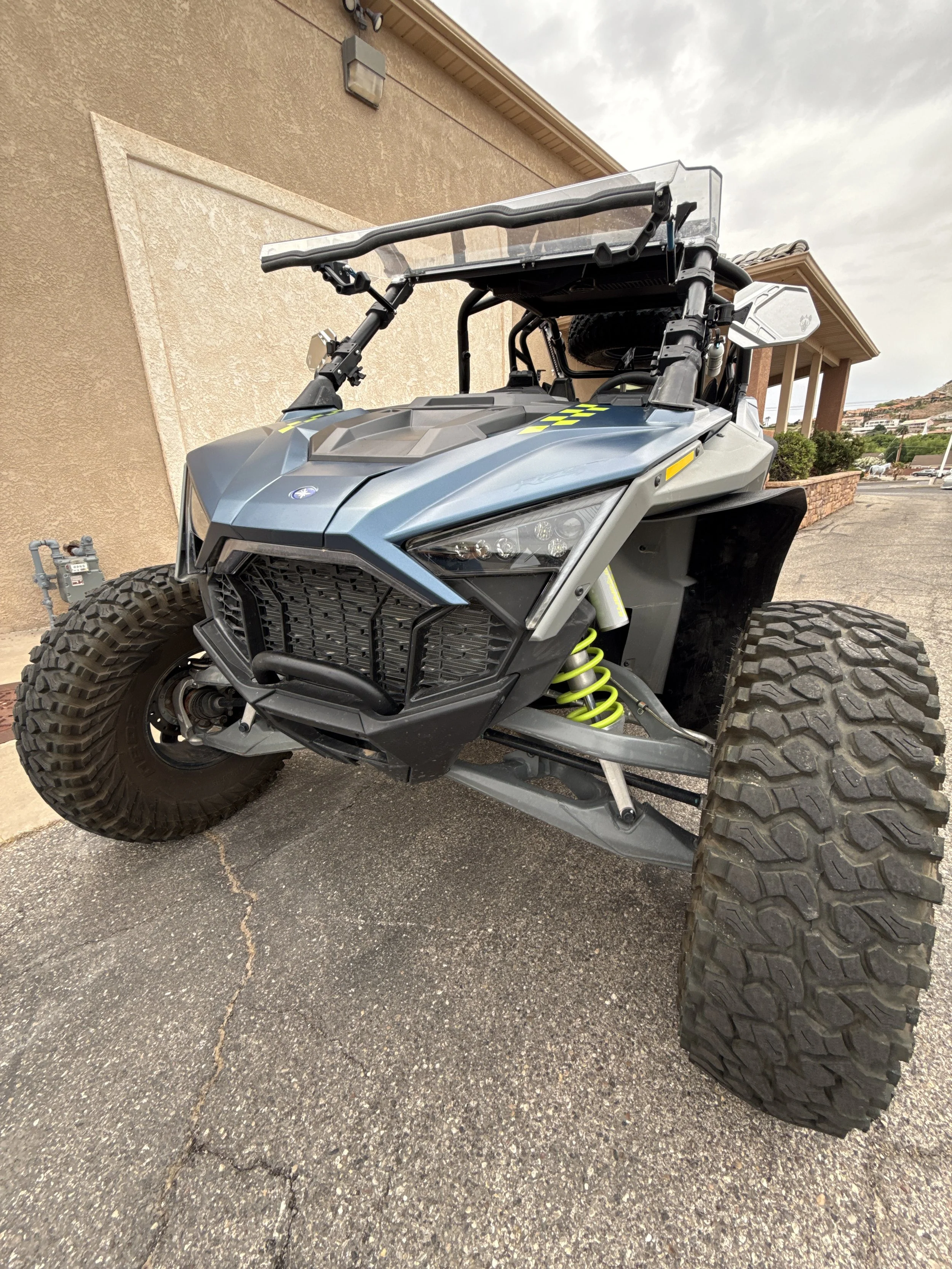 A black off-road dune buggy with large tires, exposed suspension, and a metal frame, parked on a concrete sidewalk in front of a beige wall with two flags on top.