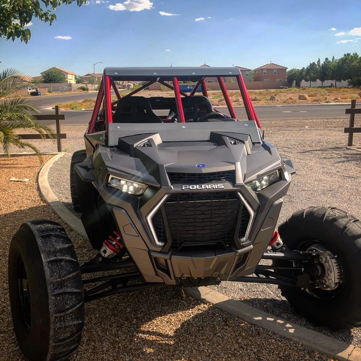 A Polaris off-road vehicle with a rugged, angular design, black and gray body, red roll cage, and large tires parked on a gravel driveway with a suburban neighborhood in the background.