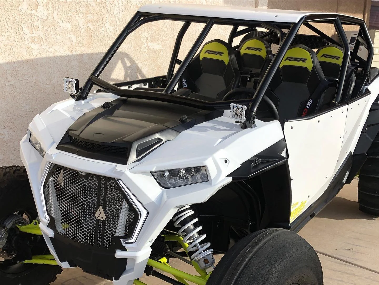 Off-road vehicle with exposed frame, four seats with 'RZR' branding, large tires, and a distinctive front grille, parked near a beige wall.