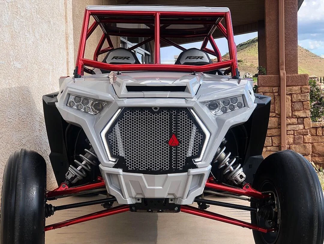 Front view of a white off-road racing vehicle with red roll cage, large black tires, and suspension system, parked on a driveway near a house.