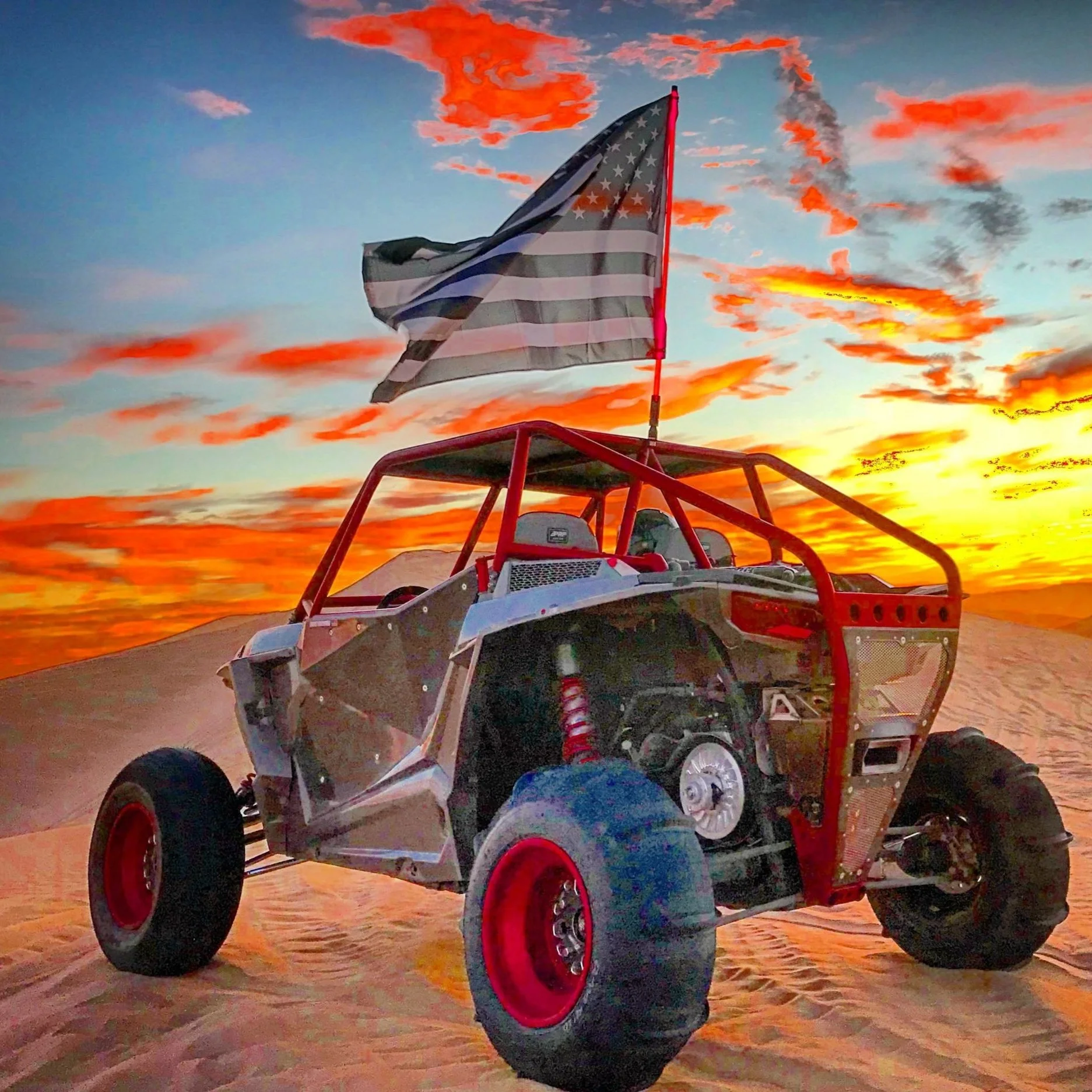 An off-road vehicle with a large American flag mounted on it, parked on sand dunes at sunset with orange and pink clouds in the sky.