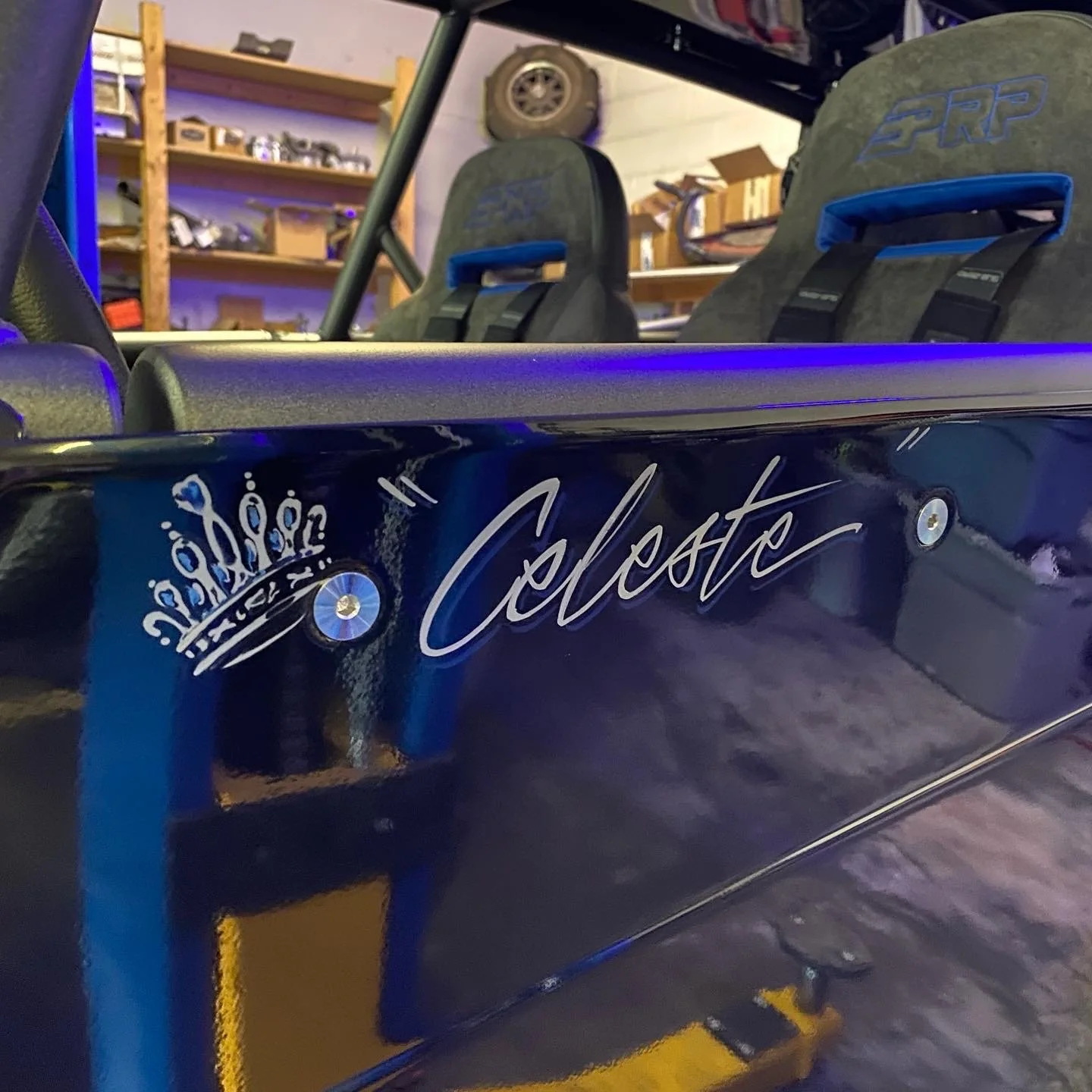 A black racing car with the name 'Celeste' written on its side and a crown logo. Two racing seats are visible inside the car. The background shows wooden shelves with various items and a tire on a wall.