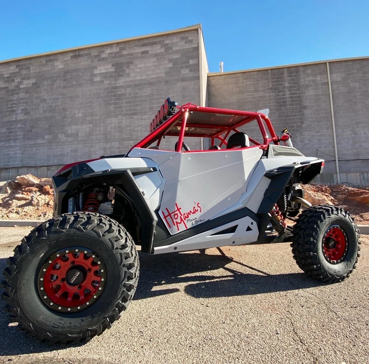 Off-road vehicle with large tires, red accents, and a gray body parked outdoors near a concrete wall on a sunny day. The vehicle has a red roll cage and a sign on the side reading 'Hot Mama's Pizza & Brew.'