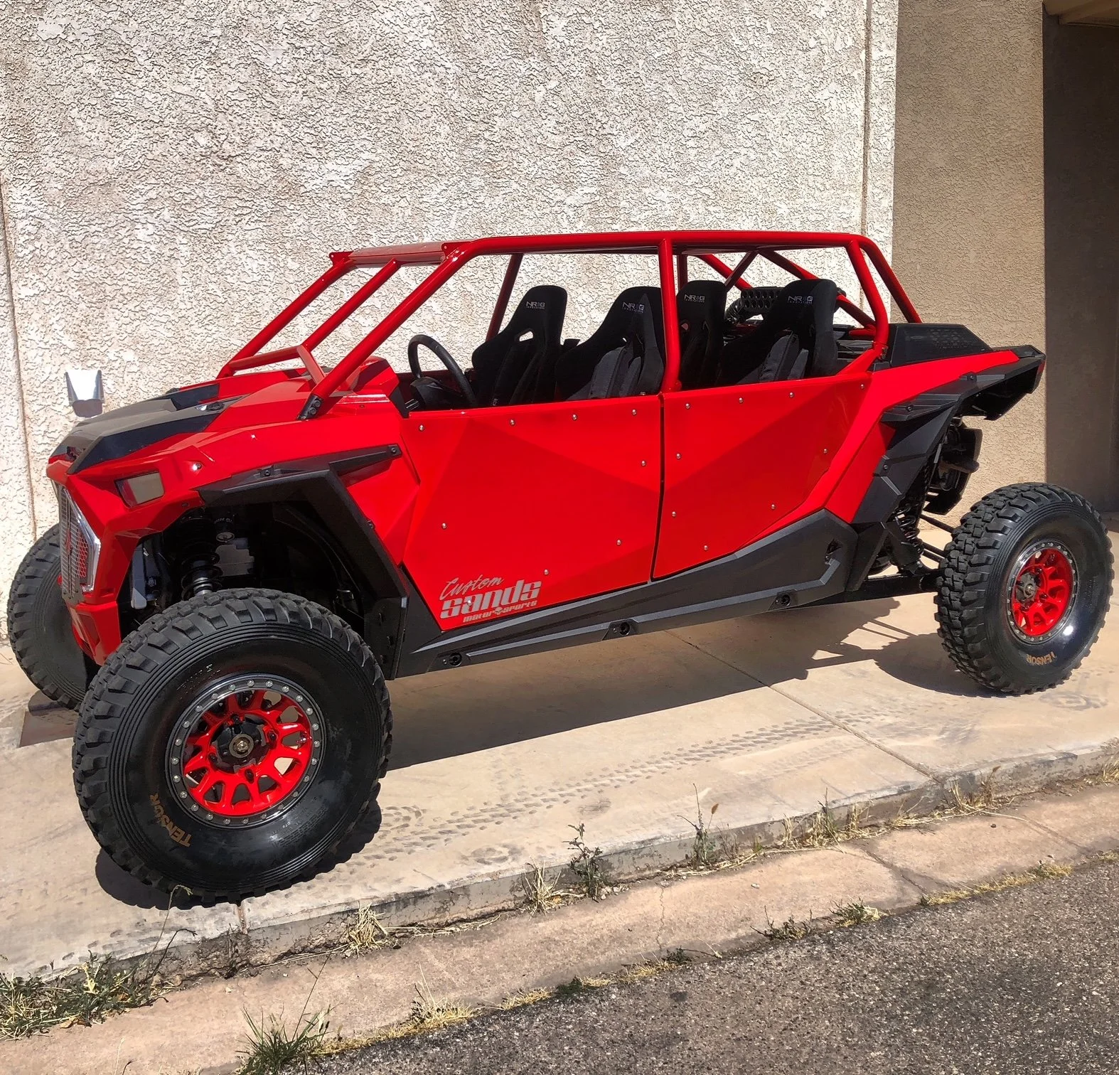 Red off-road dune buggy with black accents and four seats, parked on a concrete driveway next to a beige stucco wall.