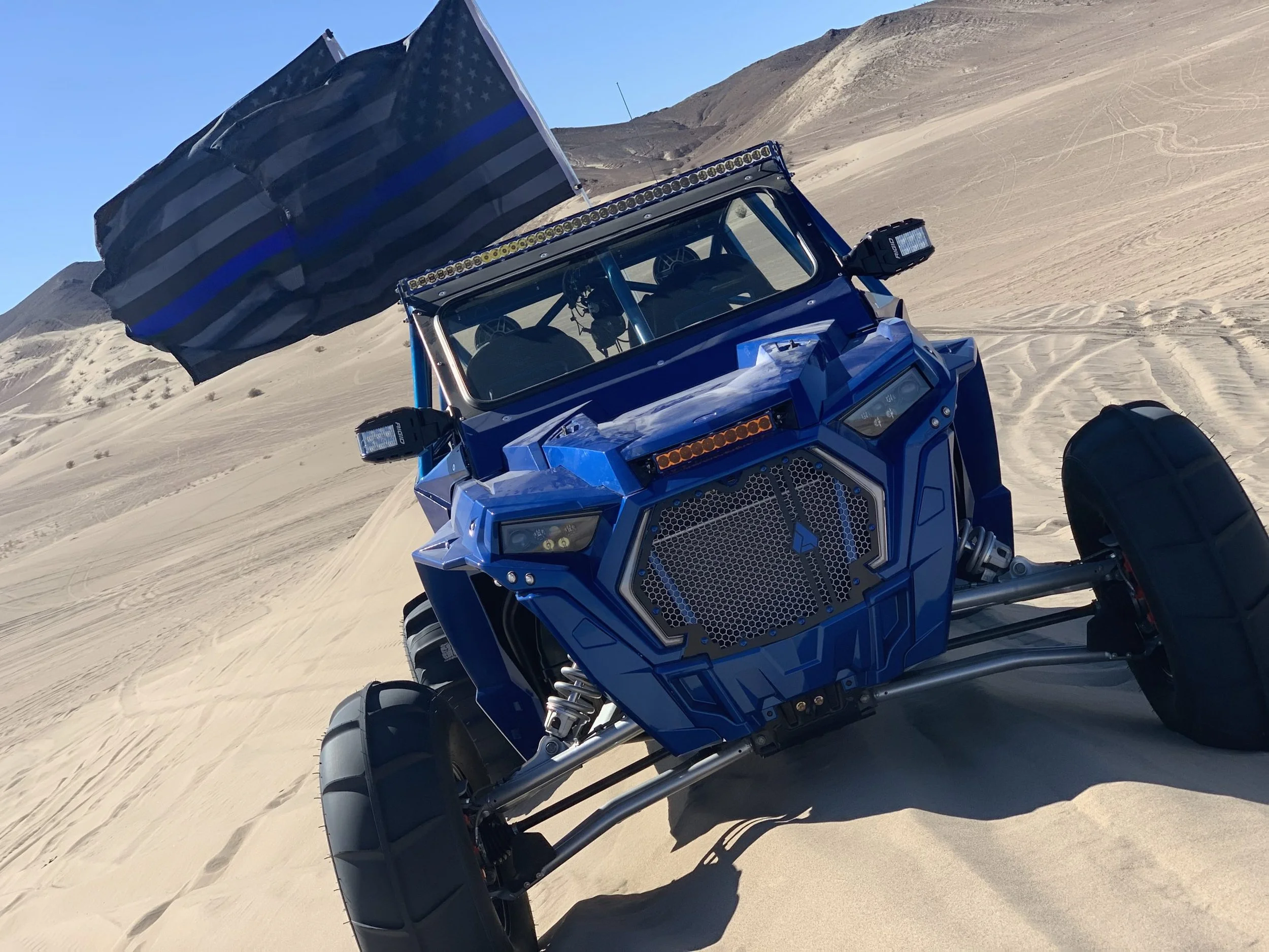 Blue off-road vehicle with a large grille and LED lights, driving on sandy dunes with a black and blue striped flag in the background.