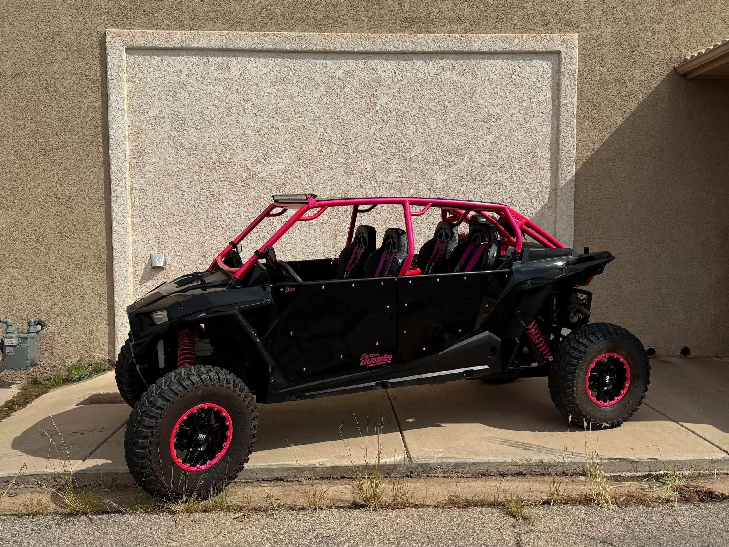 A custom off-road vehicle with black body and pink frame, featuring six seats with racing harnesses, large knobby tires with pink accents, parked on a driveway in front of a beige wall.