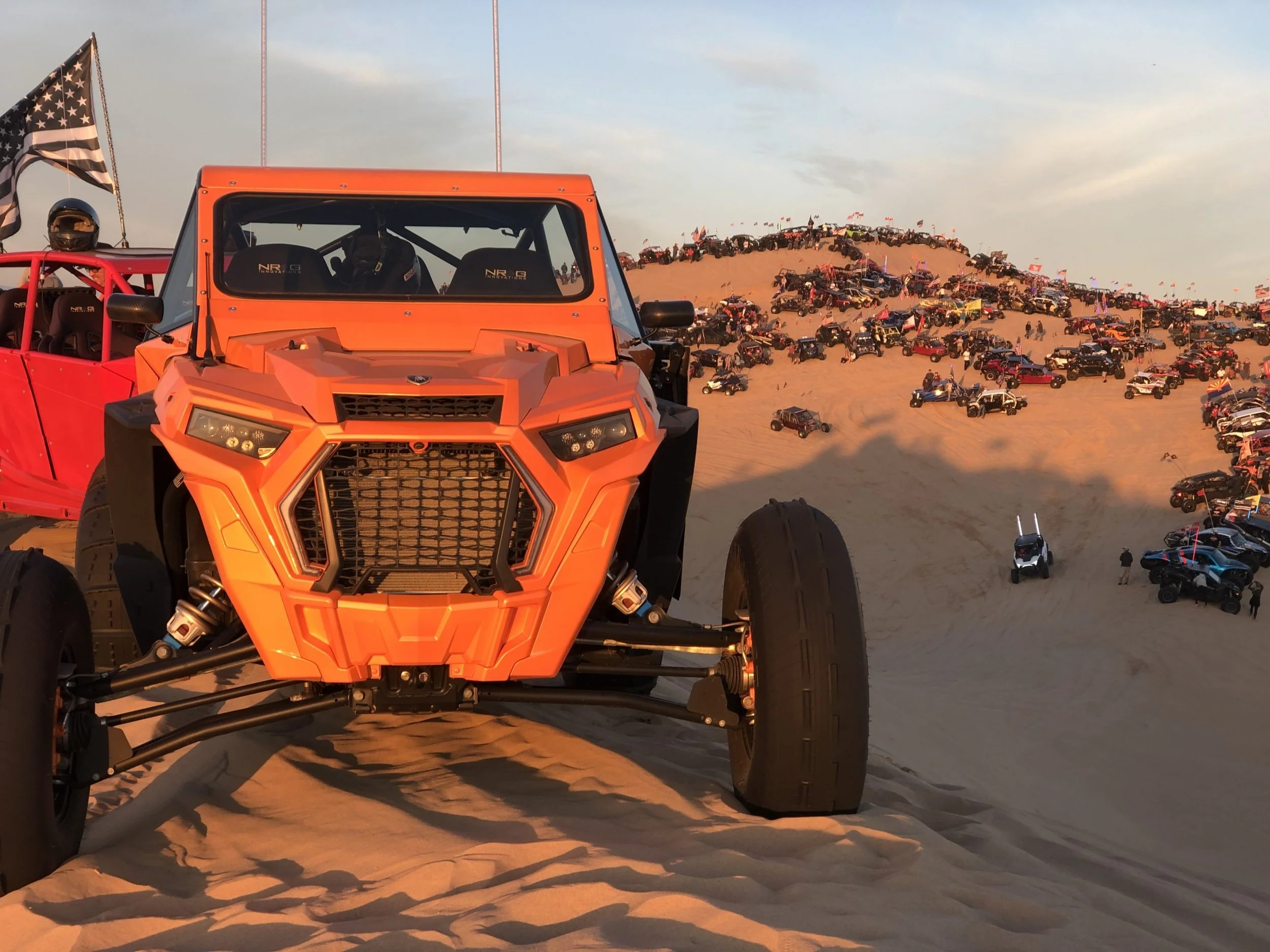 Orange off-road vehicle parked on a sandy dune with a desert landscape filled with multiple off-road vehicles and a dune in the background.
