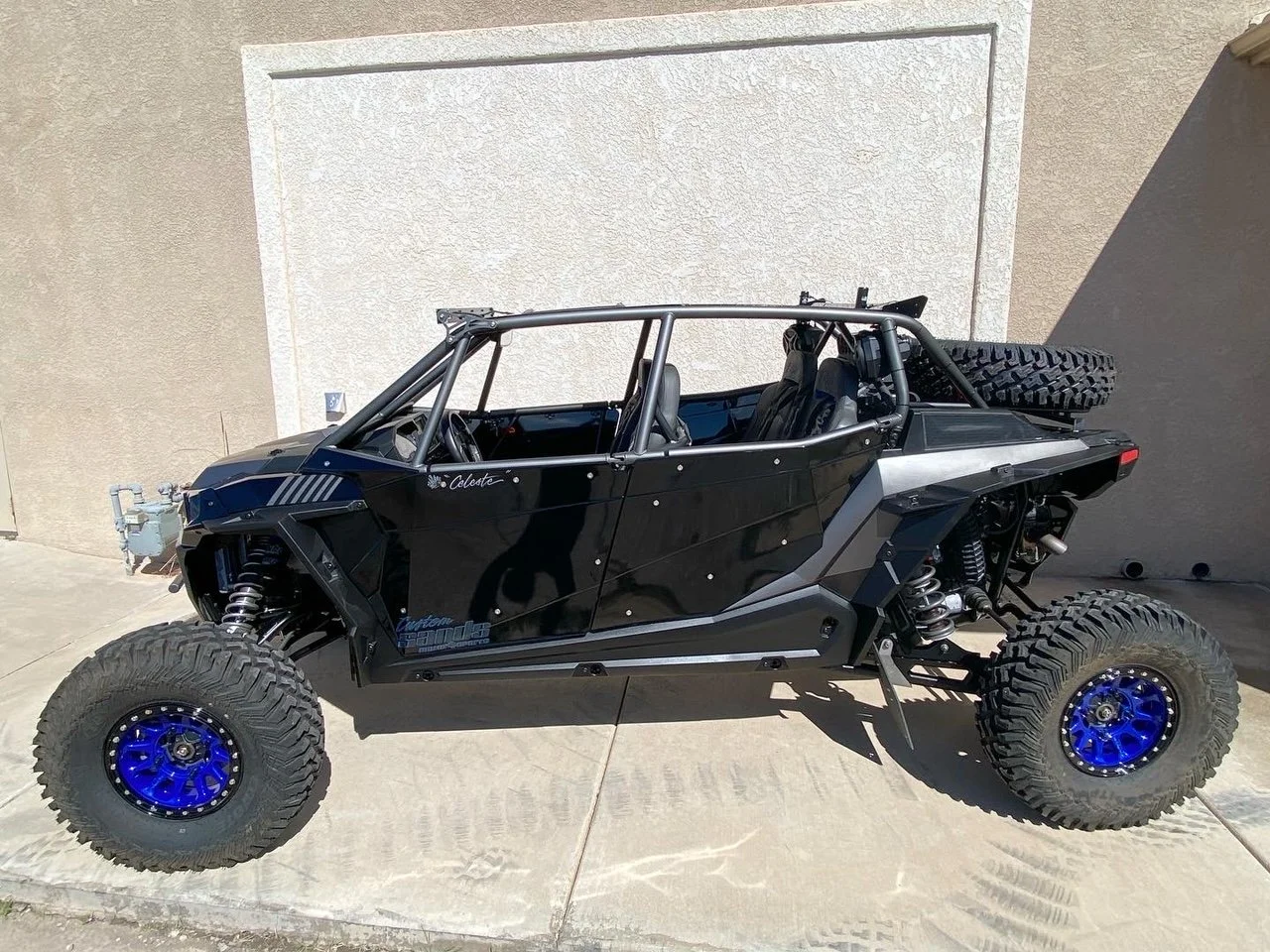 Off-road vehicle with a black body, large tires, and blue rims parked on concrete next to a beige wall.