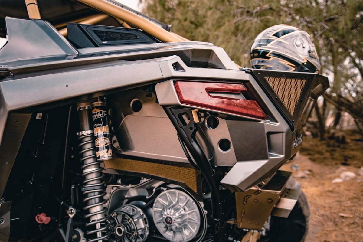 Close-up of a futuristic off-road vehicle's rear, showcasing its suspension system, engine components, and a sleek helmet resting on the vehicle.