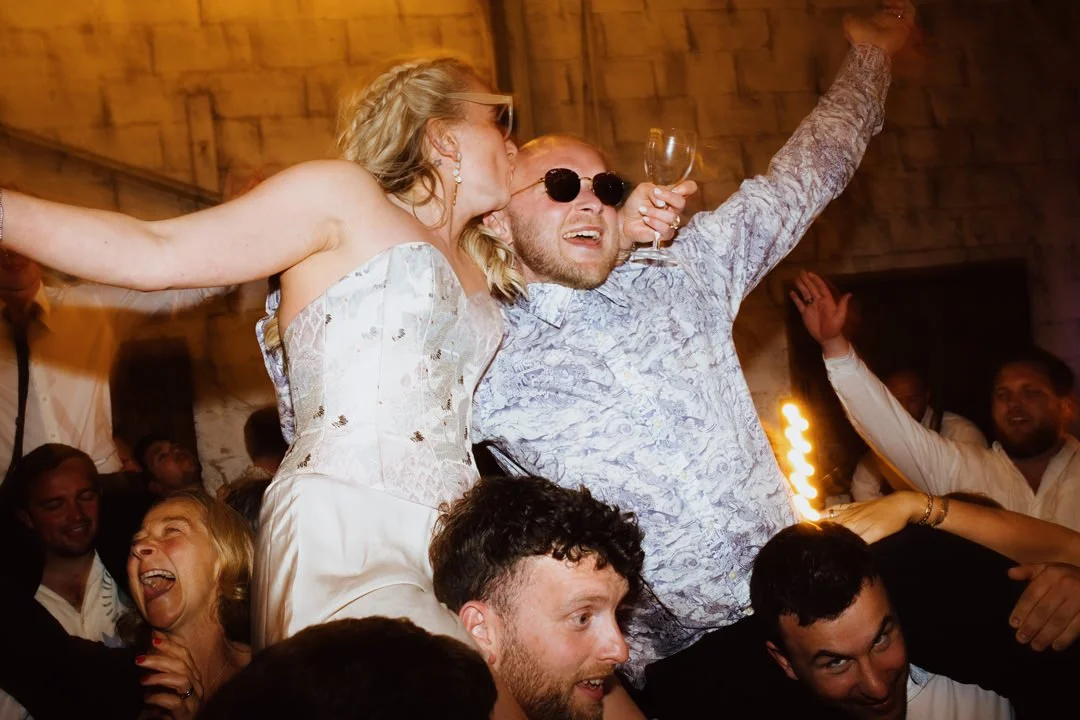 A lively celebration with people dancing and having fun at a party, with a woman kissed on the cheek and a man smiling with sunglasses, surrounded by other smiling guests.