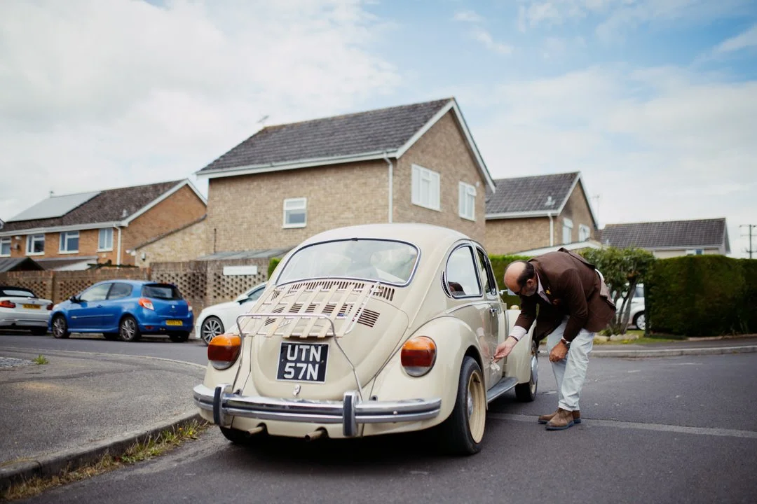 A man in a brown blazer and gray pants bending down near the front of a vintage cream-colored Volkswagen Beetle parked on a residential street.