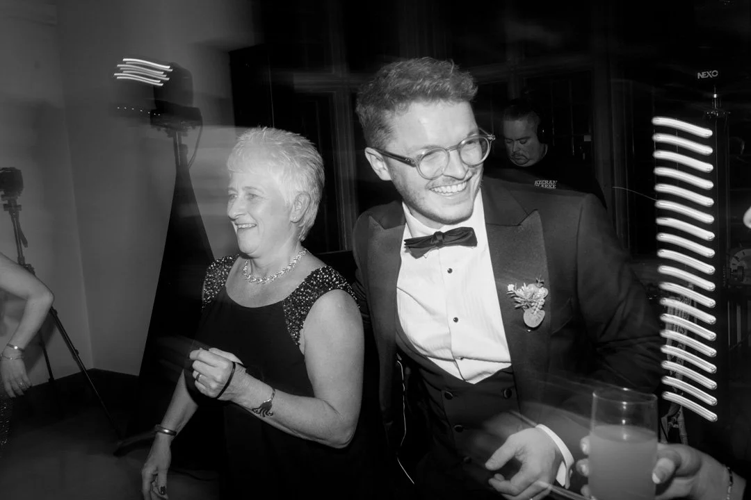 A black and white photo of a young man in glasses and a tuxedo with a bowtie, smiling and holding a drink, and an older woman with short hair, also smiling, at a formal event.