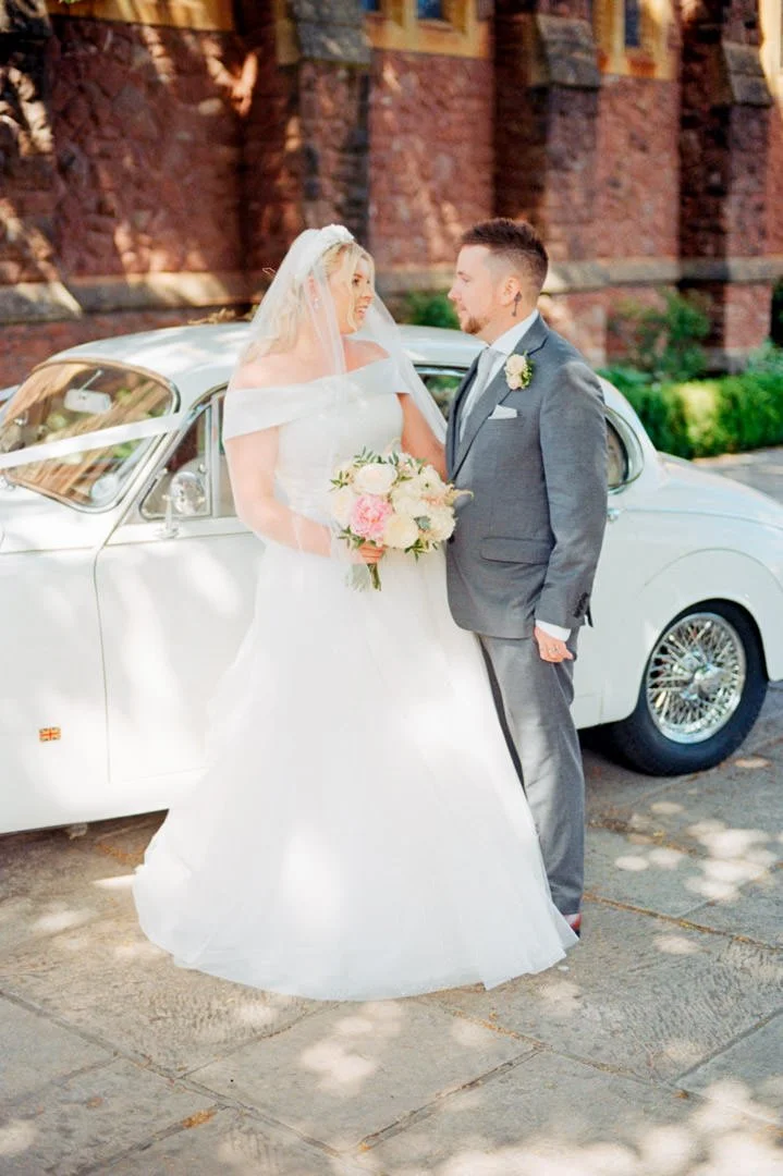 A bride and groom stand close together in front of a white vintage car, sharing a moment on their wedding day. The bride is dressed in a white wedding gown and holding a bouquet, while the groom is wearing a gray suit with a boutonniere.