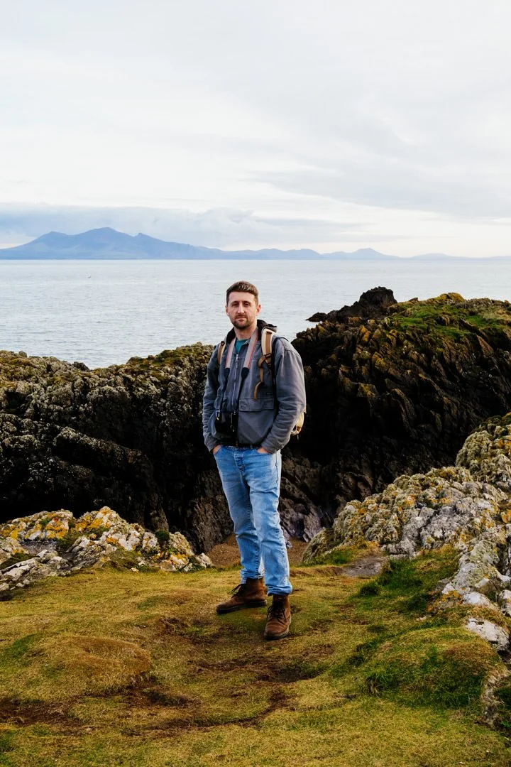 man waring blue jeans and grey jacket next to large lake. he has two cameras around his neck