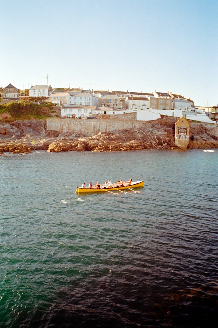 A group of people rowing a yellow boat in the water near a rocky shoreline with houses on a hill in the background.