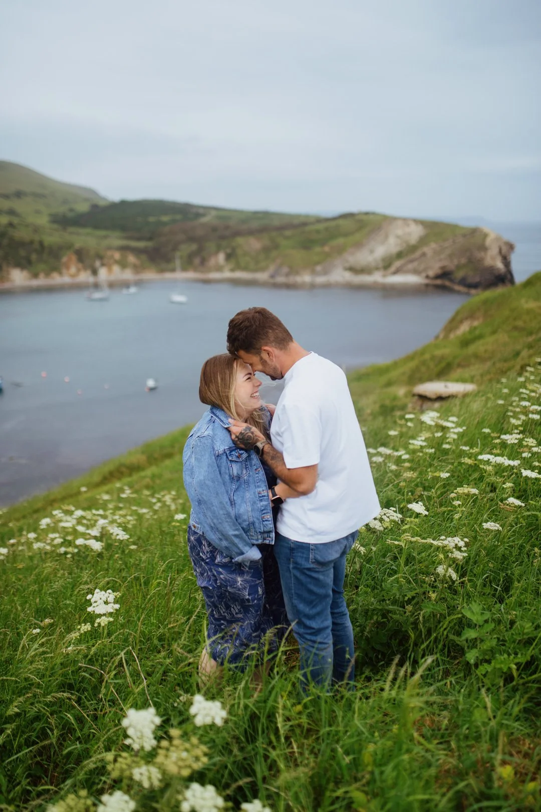A couple on a grassy hillside near a bay, embracing and smiling at each other with sailboats on the water and green hills in the background.