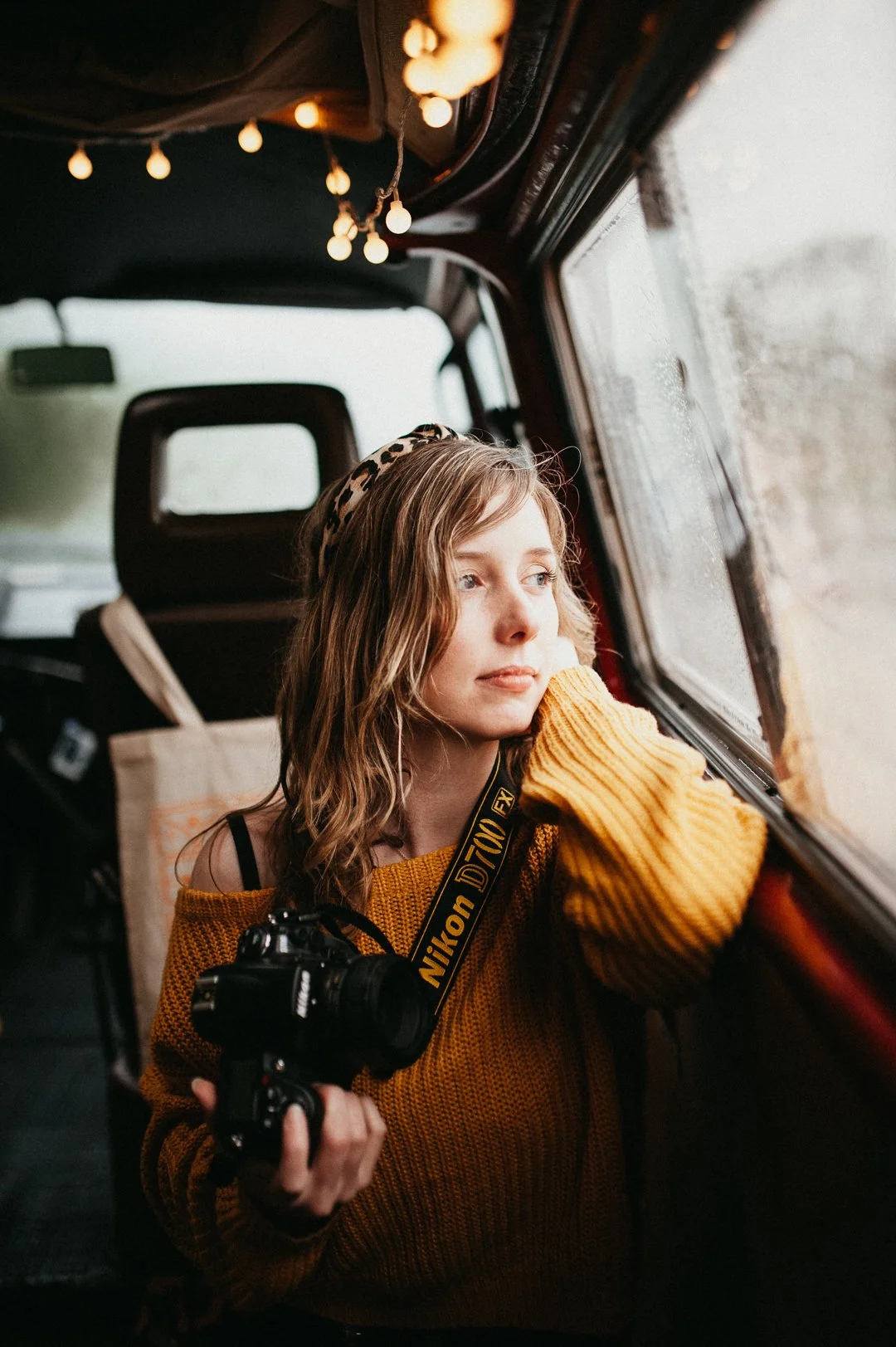 A young woman with wavy blonde hair, wearing a leopard print headband and a yellow sweater, sitting on a vehicle's seat, gazing out the rain-soaked window, holding a Nikon DSLR camera around her neck.