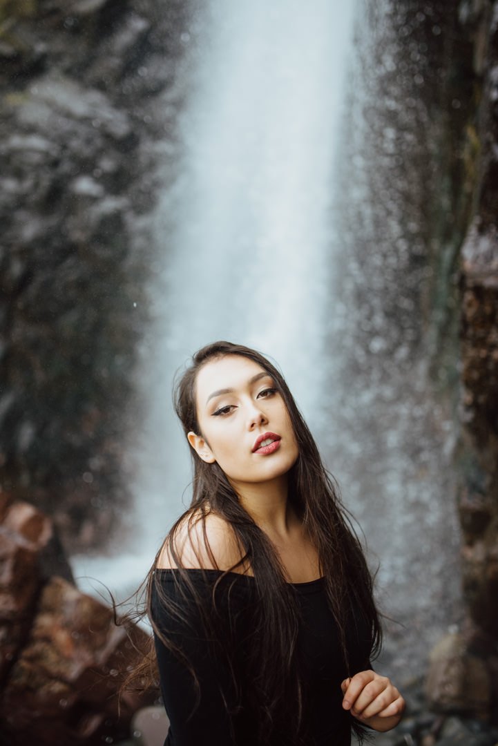 A young woman with long dark hair and makeup posing in front of a waterfall, wearing a black off-the-shoulder top.