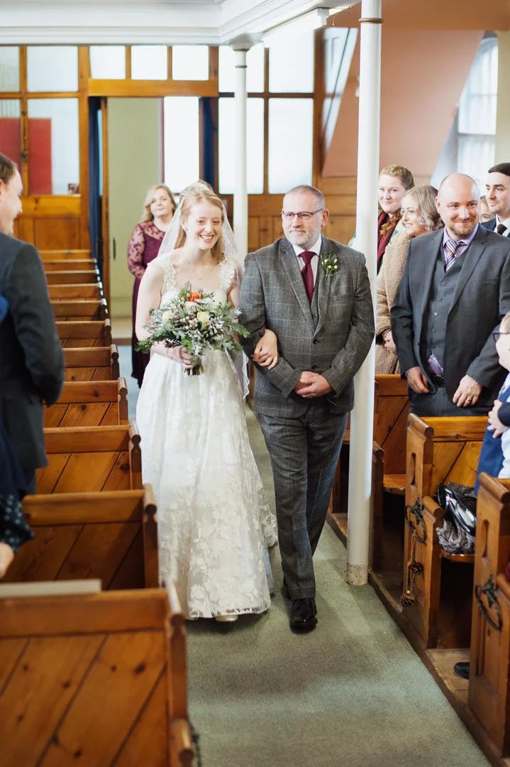A bride walking down the aisle with an escort at a wedding ceremony in a church. The bride is smiling and holding a bouquet of flowers, wearing a white lace dress. The church has wooden pews and large windows.