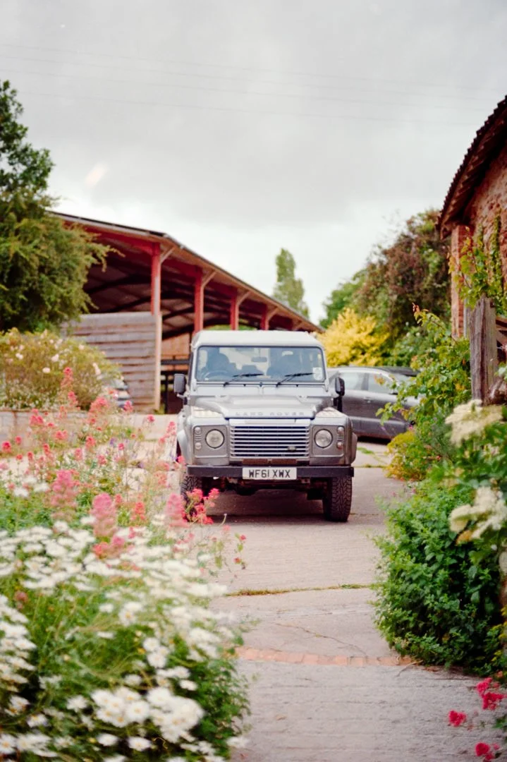 A white Land Rover Defender parked on a stone pathway surrounded by colorful flowers and greenery in a garden or courtyard setting, with a wooden building in the background.