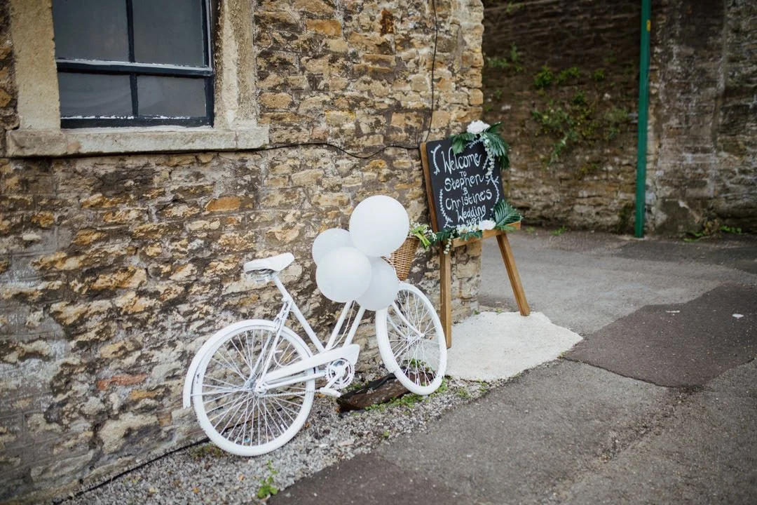 white bicycle with three white balloons on handle bars. it is next to a black wooden saying saying wedding