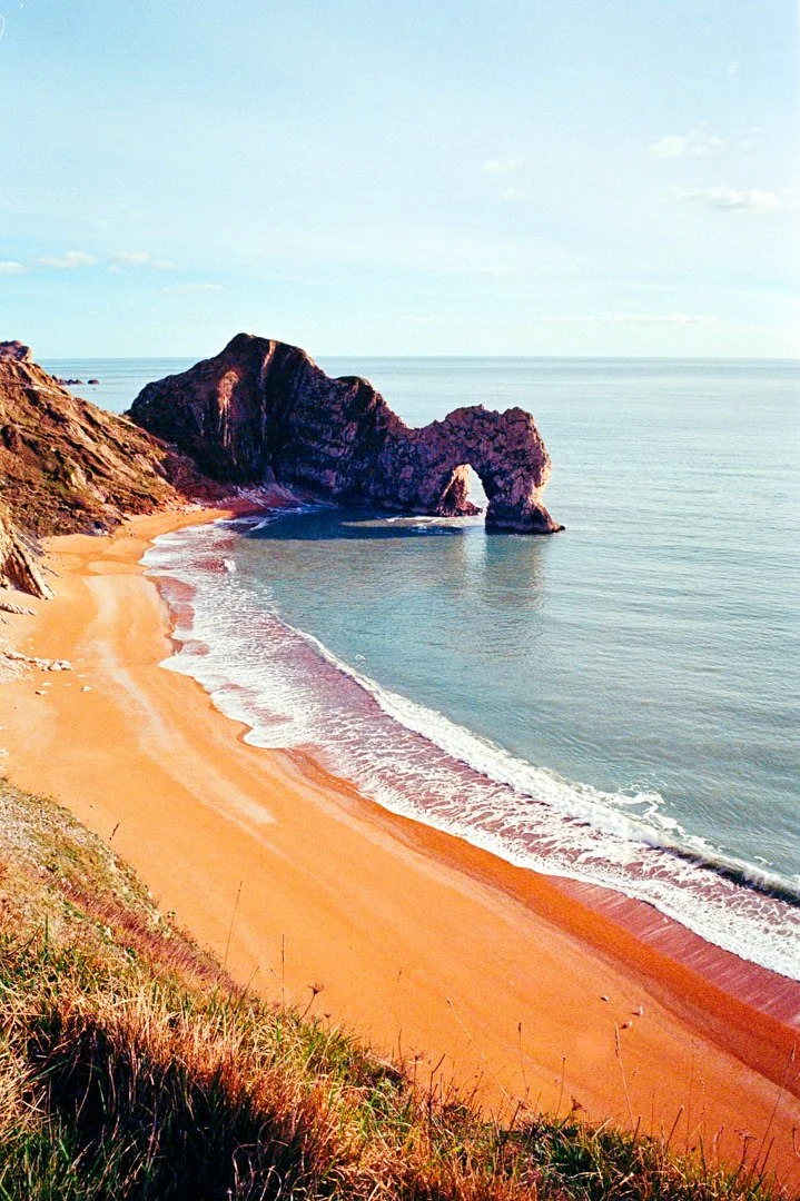 Scenic view of a sandy beach with a natural rock arch formation extending into the ocean, under a partly cloudy sky.