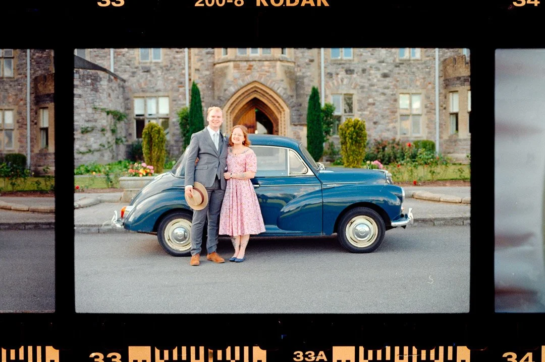 A man and woman standing in front of a vintage blue car with a stone castle-like building in the background.