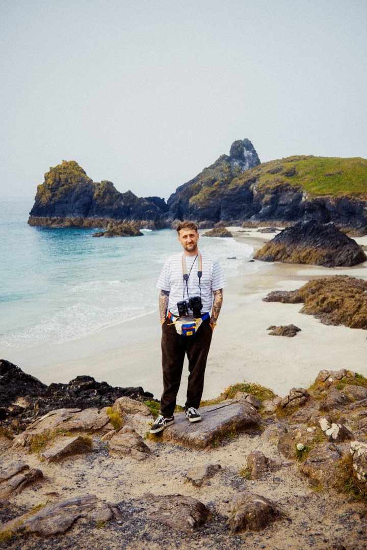 man with two old film cameras stood on cliff with rugged coast line behind, kynance cove