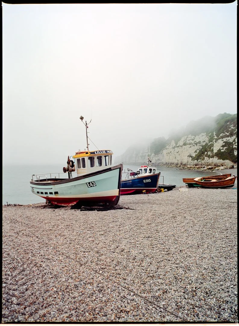 small blue and red boats on the shingle beach under foggy sky, there is a cliff in backround, Kodak portra 160 film