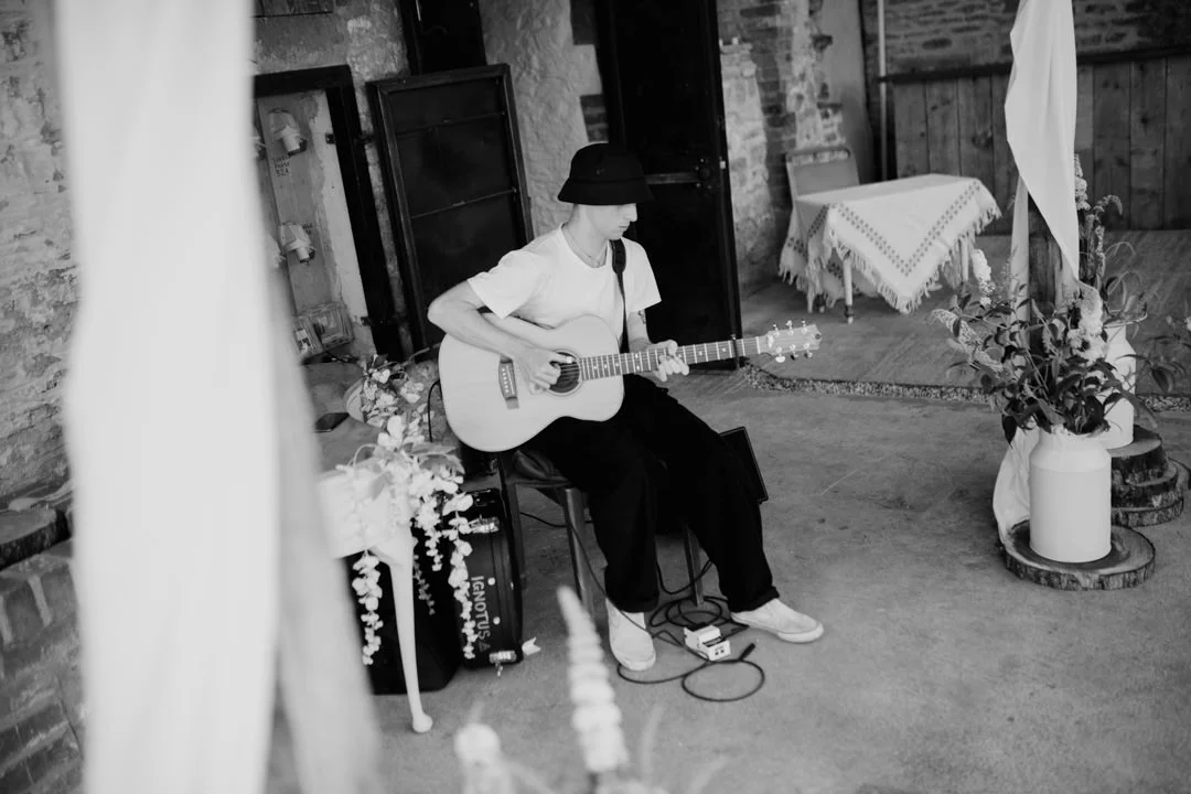 man waring black hat playing a guitar. he is sat on a stool next to a large speaker