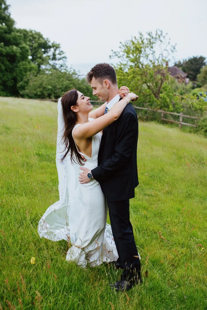 A bride and groom embracing and smiling at each other in a grassy field with trees and a house in the background.