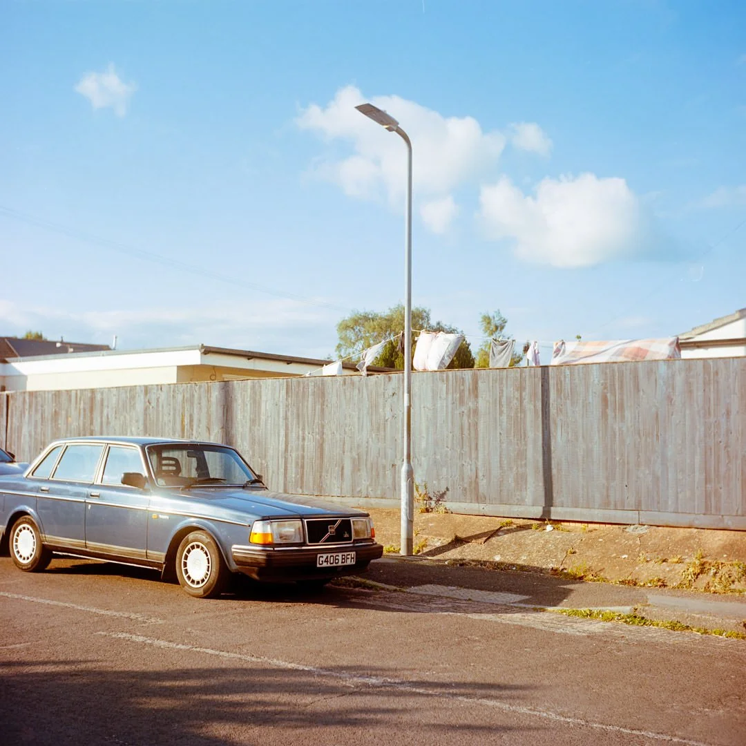 A vintage blue station wagon parked on a street next to a wooden fence with laundry hanging to dry on top, under a sunny sky with a few clouds.