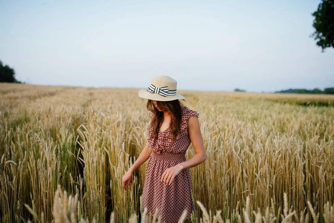 A woman in a patterned dress and wide-brimmed hat walking through a wheat field.