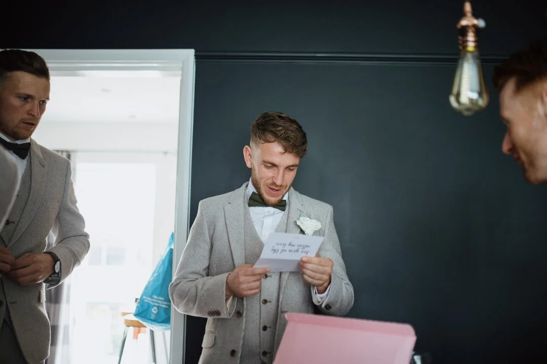 wedding groom reading a letter stood at a large table. he is waiting a grey suit and waistcoat