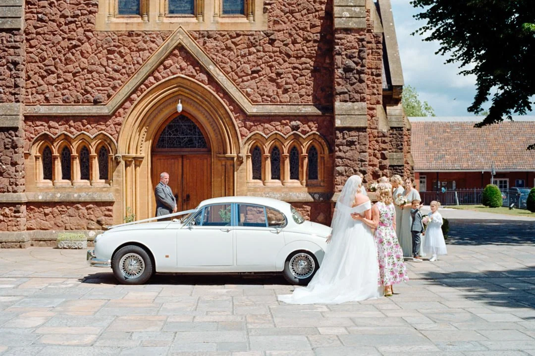 A bride and groom in wedding attire sharing a kiss in front of a white vintage car parked outside a red brick church, with wedding guests and a man standing near the church door.