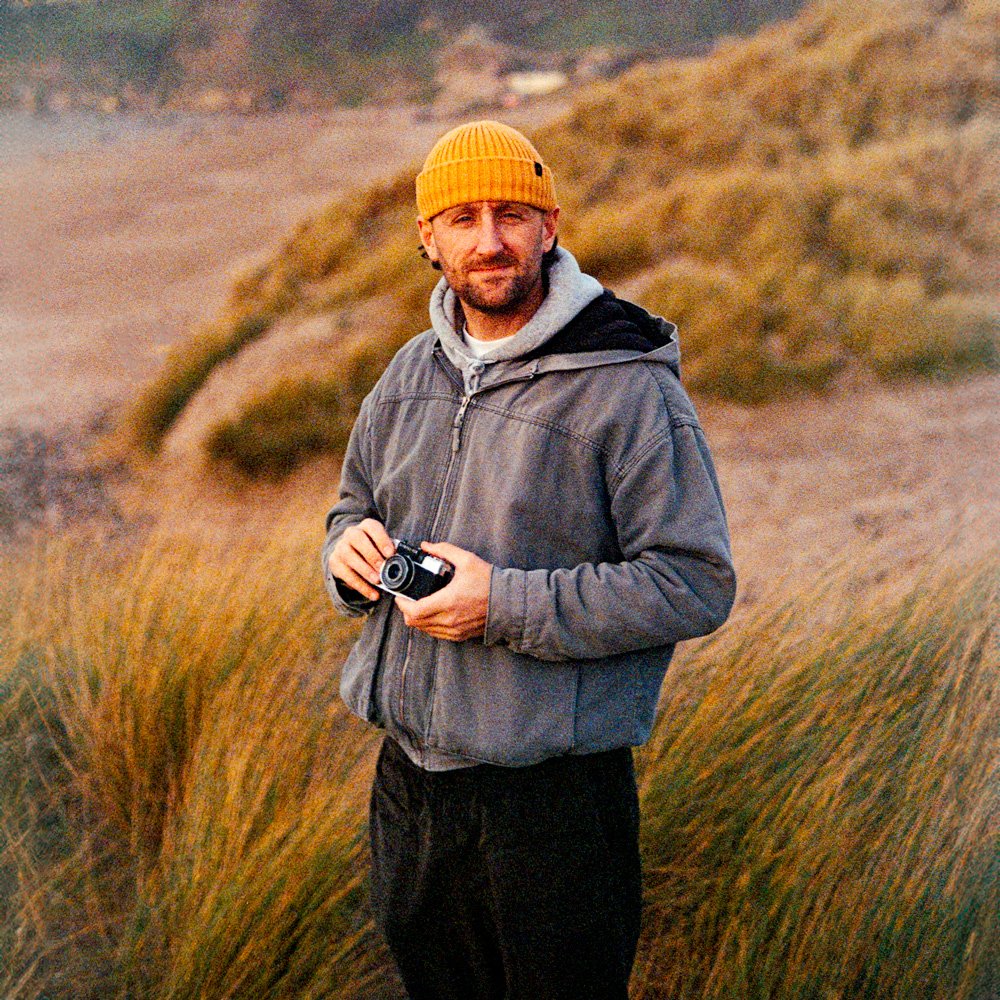 man on sand dunes holding film camera waring yellow beanie hat