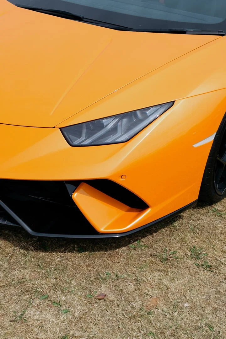 Close-up of the front part of an orange sports car, showing the headlight, part of the hood, and an air intake vent.