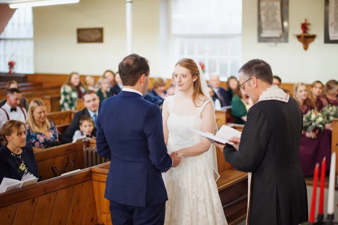 A bride and groom standing at the altar in a church during their wedding ceremony, with an officiant reading from a book.