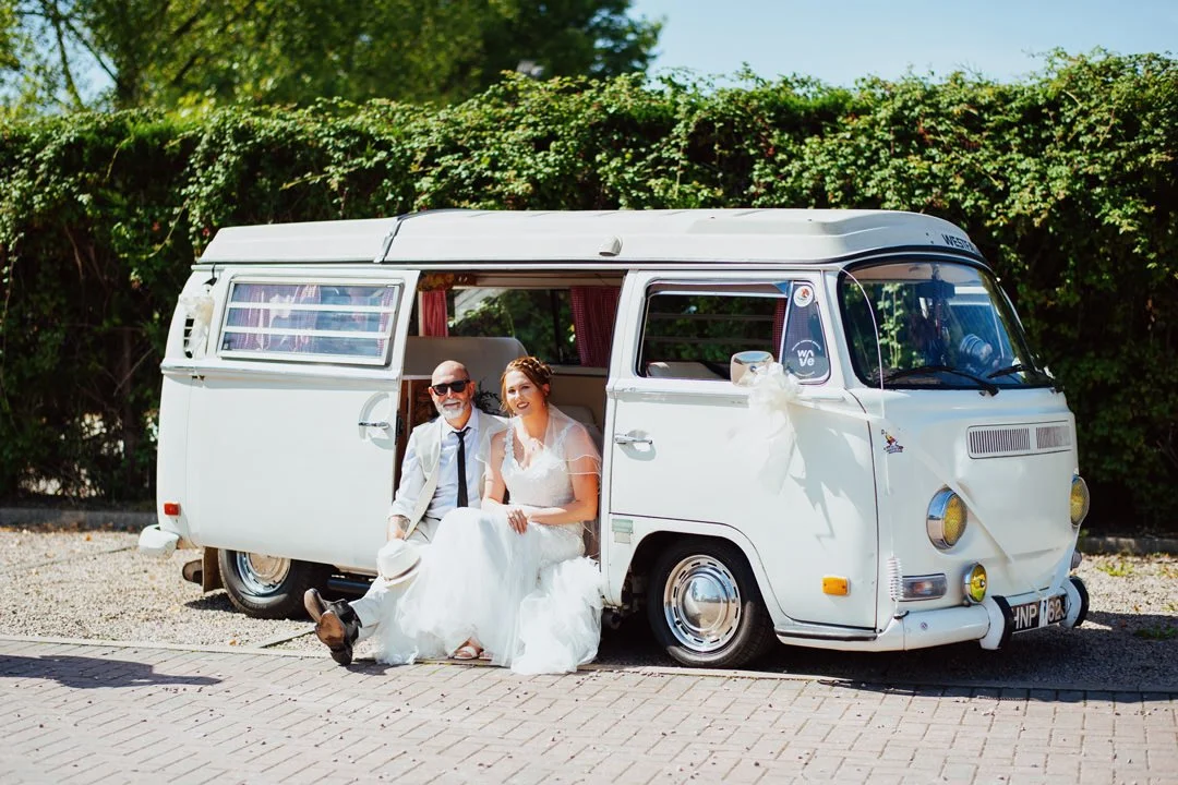 A bride and groom sitting on the step of a white vintage van decorated with a white bow, parked outdoors on a sunny day.