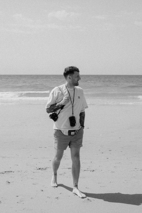 man walking on beach carrying two film cameras, black and white photo