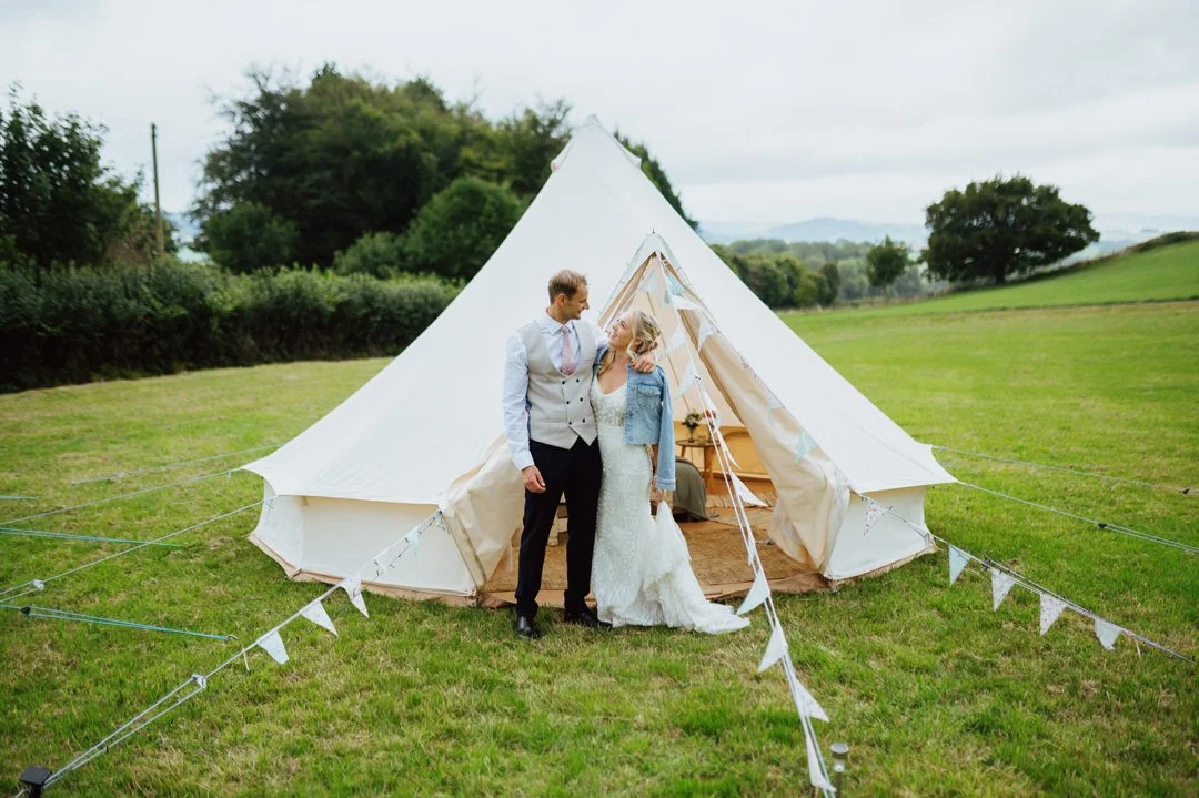 A couple dressed in wedding attire standing outside a large white tent in a grassy field, with a backdrop of trees and open sky.