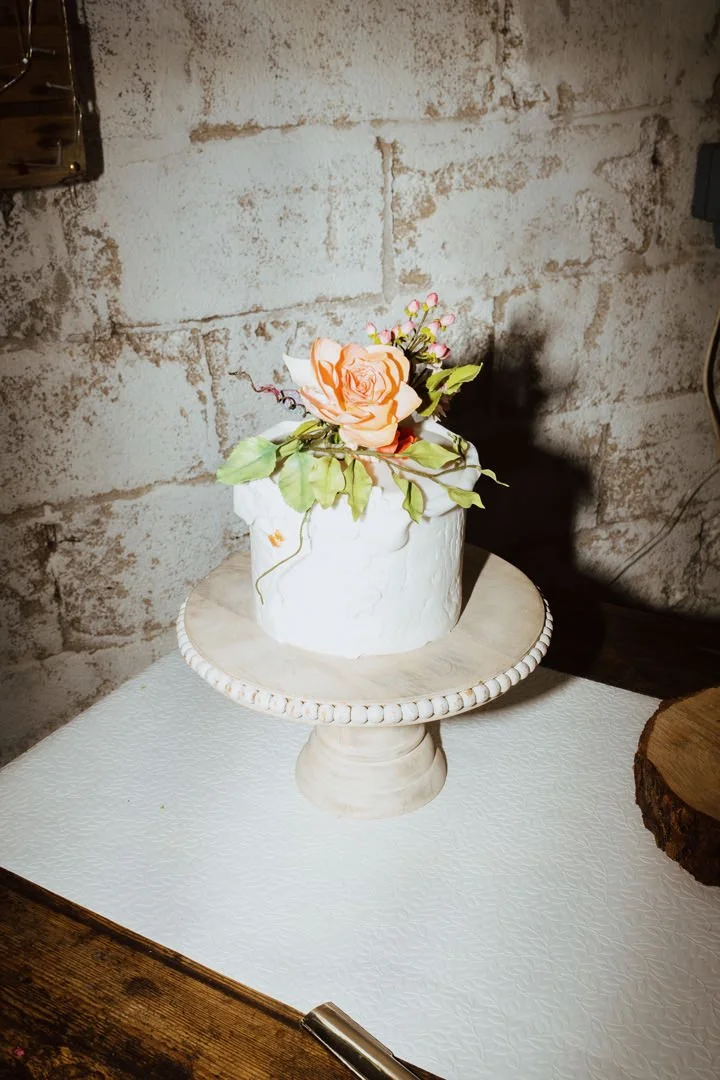 A small white cake with a textured icing top on a pedestal cake stand, decorated with a peach-colored rose, pink flowers, and green leaves, placed against a brick wall background.